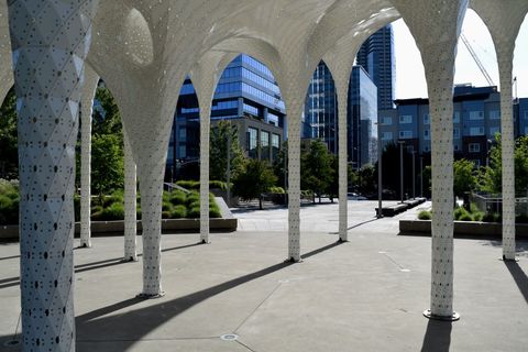 In the foreground, there is a plaza with greenery and benches. In the background, the fluted columns of the sculpture Piloti rise up to form an undulating canopy.