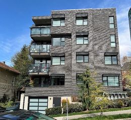 A picture containing an apartment building, outdoor space, trees, sky and the roof of a car parked on the street. The apartment building features five levels with four visible balconies and a street level garage door. 