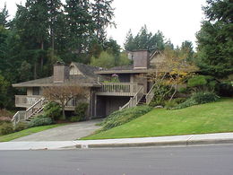 Photo shows multifamily housing in a stacked style with units over garages and stacked with shared walls. The photo shows dwelling units over garage with access via stairs. The photo also shows a driveway, sidewalk and grass with greenery and trees behind the multifamily housing. This photo is provided as an example of a multifamily dwelling type. 